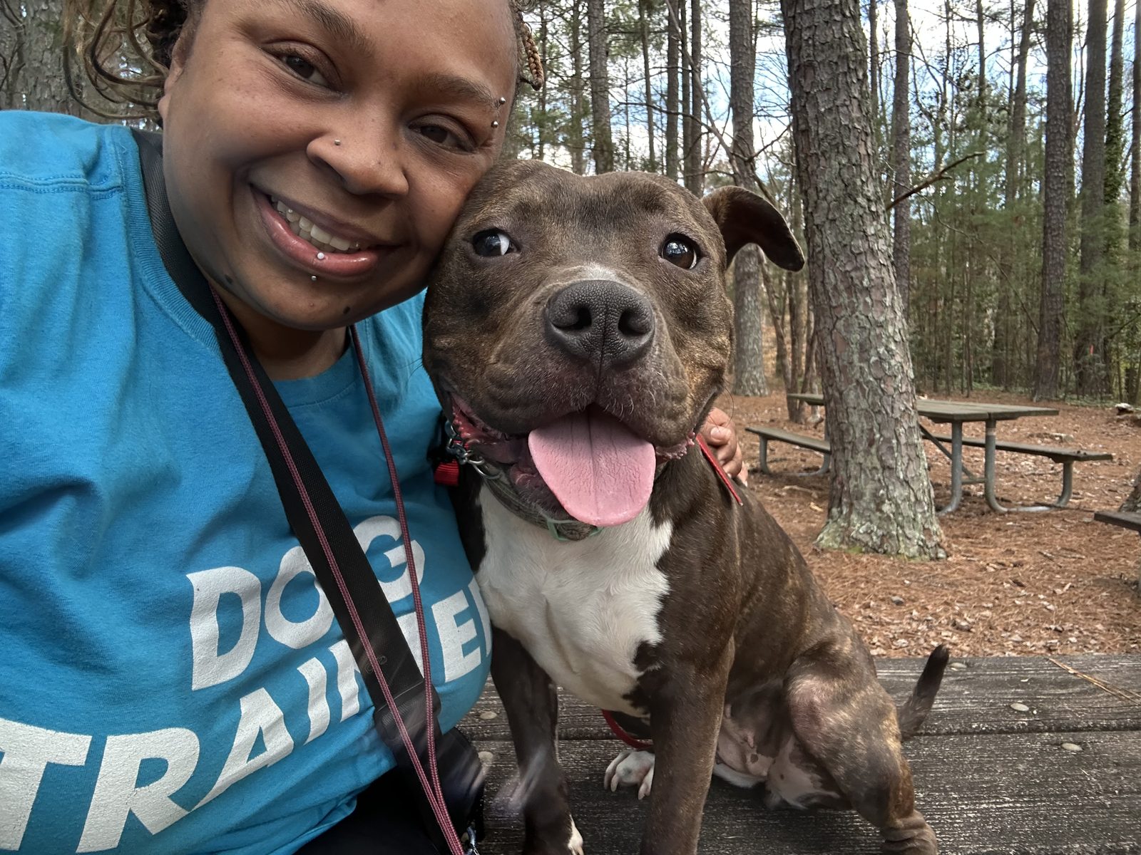 Jen smiling cheek to cheek with a brindle pit bull in a pine forest